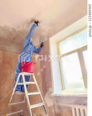 A worker dressed in a blue suit skillfully washes chalk and lime off the ceiling while balancing on a ladder positioned near a window 123469365