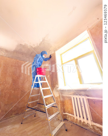 A worker dressed in a blue suit skillfully washes chalk and lime off the ceiling while balancing on a ladder positioned near a window A worker dressed in a blue suit skillfully washes chalk and lime off the ceiling while balancing on a ladder positioned near a window 123469379