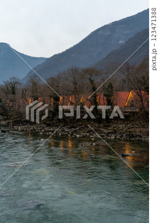 River Lodges Mountains Dusk: Illuminated riverside lodges nestled in mountainous terrain at twilight, offering tranquil getaway. 123471388