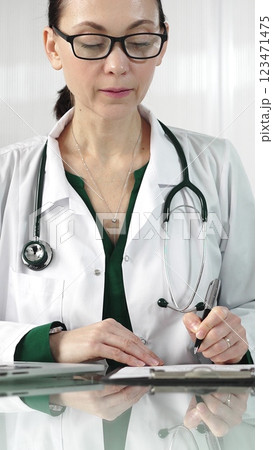 Female doctor in a green blouse under a medical coat is filling up medication history records on a glass desk in the clinic, vertical portrait view. Medicine concept 123471475