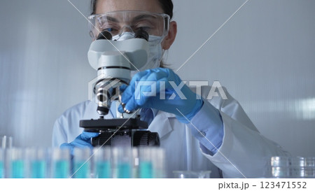 Scientist woman, wearing a lab coat, mask, blue gloves and safety glasses, is adjusting a microscope surrounded by test tubes in laboratory. Medicine and science concept 123471552