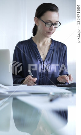 Professional woman accountant in glasses using a calculator and taking notes at a glass desk in the office. Audit and taxes in business Professional woman accountant in glasses using a calculator and taking notes at a glass desk in the office. Audit and taxes in business 123471604
