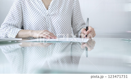 Businesswoman in white dotted blouse is signing a contract at a sleek glass desk in a modern office, showcasing professionalism and focus in her work environment. Business people concept Businesswoman in white dotted blouse is signing a contract at a sleek glass desk in a modern office, showcasing professionalism and focus in her work environment. Business people concept 123471731