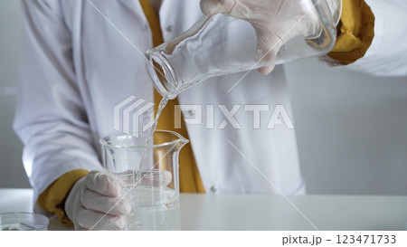Unknown scientist wearing a lab coat and gloves is carefully pouring a liquid from one beaker into another, close up. Science and medicine concepts Unknown scientist wearing a lab coat and gloves is carefully pouring a liquid from one beaker into another, close up. Science and medicine concepts 123471733