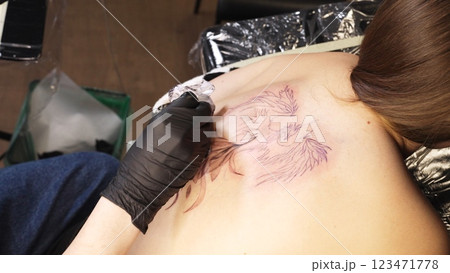 Professional tattoo artist wearing black gloves drawing intricate design on woman's back while working in sleek, well lit tattoo studio with modern equipment Professional tattoo artist wearing black gloves drawing intricate design on woman's back while working in sleek, well lit tattoo studio with modern equipment 123471778