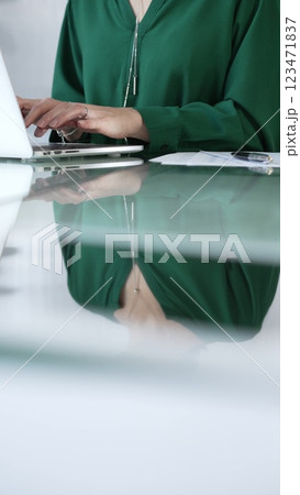 Businesswoman, wearing green blouse, is typing on laptop keyboard, preparing taxes and audit reports, with documents on glass office desk, vertical closeup view. Taxes, audit in business 123471837