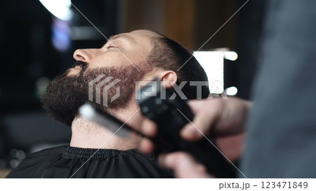 Professional barber trimming client's beard with electric razor, delivering precise styling within contemporary grooming salon setting 123471849