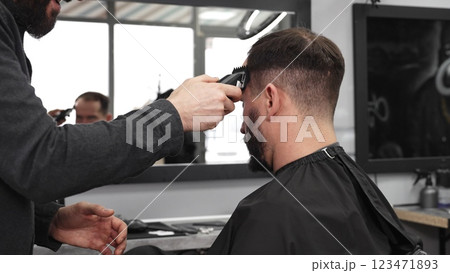Skilled barber trimming client's hair using electric razor, creating contemporary hairstyle in modern barbershop setting with precise, professional technique Skilled barber trimming client's hair using electric razor, creating contemporary hairstyle in modern barbershop setting with precise, professional technique 123471893