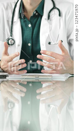 Doctor man wearing a stethoscope and lab coat is gesturing with hands while explaining a diagnosis to a patient across a glass desk with a laptop. Medicine concept 123471970