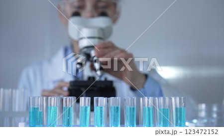 Laboratory tubes, filled with a blue liquid, are in focus, closeup view. Female scientist researcher is using a microscope at the background Laboratory tubes, filled with a blue liquid, are in focus, closeup view. Female scientist researcher is using a microscope at the background 123472152