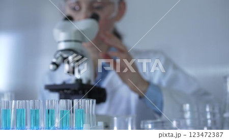 A female scientist, wearing a lab coat and safety glasses, is adjusting a binocular microscope surrounded by test tubes in laboratory setting. Medicine and science A female scientist, wearing a lab coat and safety glasses, is adjusting a binocular microscope surrounded by test tubes in laboratory setting. Medicine and science 123472387