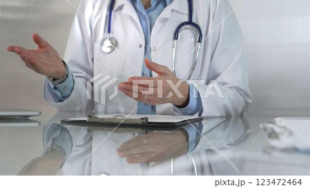 Doctor woman wearing a blue blouse, stethoscope and lab coat is gesturing with hands while explaining a diagnosis to a patient across a glass desk with a laptop. Medicine concept Doctor woman wearing a blue blouse, stethoscope and lab coat is gesturing with hands while explaining a diagnosis to a patient across a glass desk with a laptop. Medicine concept 123472464