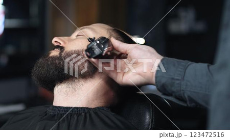 Professional barber trimming client's beard with electric razor, delivering precise styling within contemporary grooming salon setting Professional barber trimming client's beard with electric razor, delivering precise styling within contemporary grooming salon setting 123472516