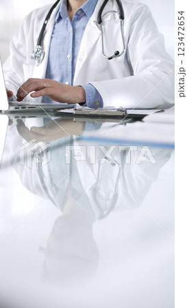Unrecognizable doctor man in a blue shirt under a medical coat is using technology in medicine. Close-up of hands typing on a laptop on a glass desk in clinic, vertical view. Health care theme concept Unrecognizable doctor man in a blue shirt under a medical coat is using technology in medicine. Close-up of hands typing on a laptop on a glass desk in clinic, vertical view. Health care theme concept 123472654