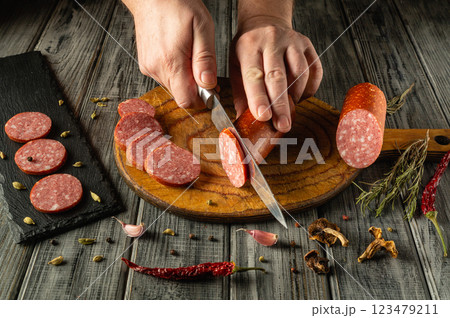 A pair of hands skillfully slicing salami on a wooden cutting board. Surrounding the meat are various spices and herbs, adding a rustic touch to the kitchen atmosphere A pair of hands skillfully slicing salami on a wooden cutting board. Surrounding the meat are various spices and herbs, adding a rustic touch to the kitchen atmosphere 123479211