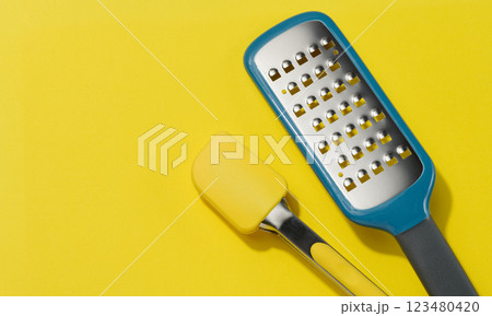Yellow silicone kitchen tongs and metal vegetable grater on yellow background 123480420