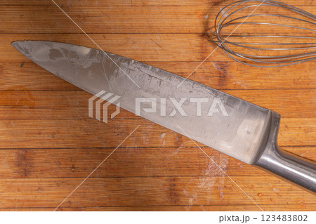 A large kitchen knife and a metal whisk placed on a wooden cutting board. 123483802