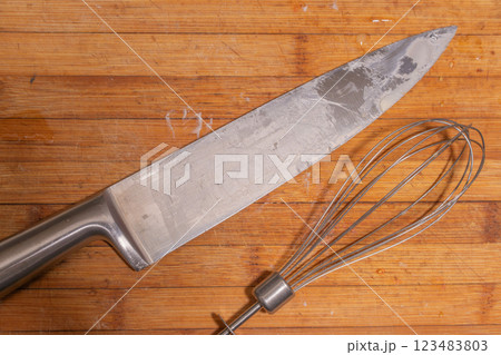 A large kitchen knife and a metal whisk placed on a wooden cutting board. 123483803