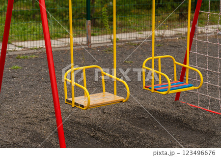 Two yellow swings with red supports stand still on a playground. The ground is made of gravel. There is a net fence behind the swings and green trees in the background 123496676