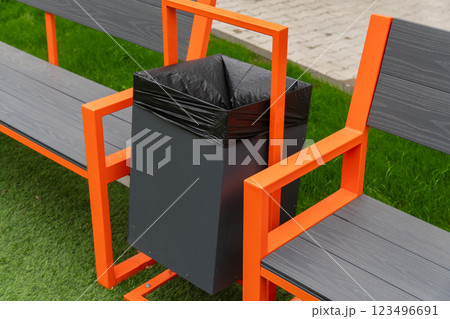 A black garbage can with a black plastic liner is mounted to an orange bench. The bench is made of metal with a gray wooden top. The bench is situated on a green lawn A black garbage can with a black plastic liner is mounted to an orange bench. The bench is made of metal with a gray wooden top. The bench is situated on a green lawn 123496691
