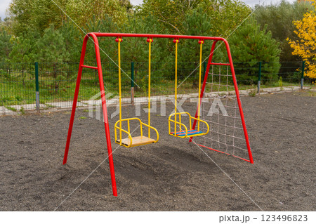 Two brightly colored swings sit in a park with soft ground cover, surrounded by lush greenery, perfect for children to play and enjoy a sunny day outdoors Two brightly colored swings sit in a park with soft ground cover, surrounded by lush greenery, perfect for children to play and enjoy a sunny day outdoors 123496823