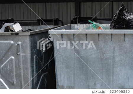 Two metal garbage containers filled with various types of rubbish are situated in a covered public space, collecting waste during daylight hours. The area shows signs of urban disarray 123496830