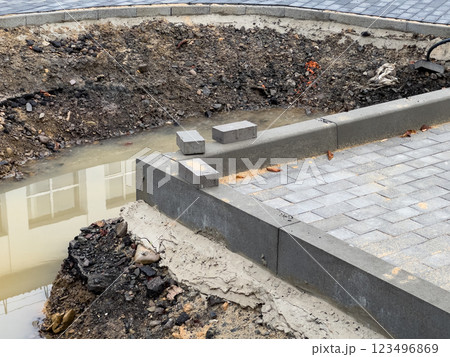 An unfinished brick pavement stretches alongside a sidewalk, featuring a trench filled with water. Loose soil and uninstalled pavers indicate ongoing construction activity 123496869