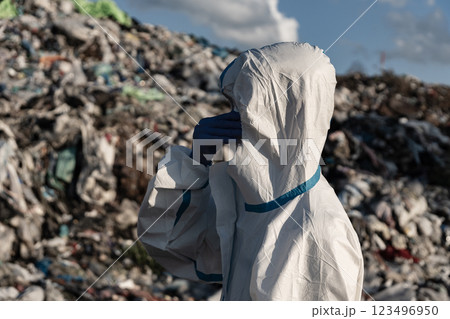 A worker clad in a protective suit inspects a sprawling landfill, surrounded by piles of waste under a partly cloudy sky. The significance of environmental safety is evident 123496950