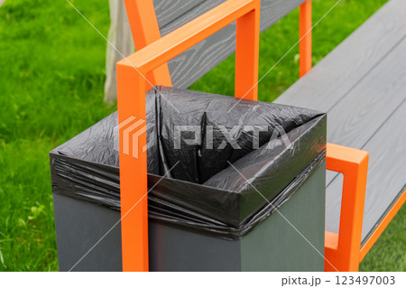 In a lush green park, a contemporary bench with orange accents showcases an attached trash bin, inviting visitors to enjoy the outdoors while keeping the area clean and tidy 123497003