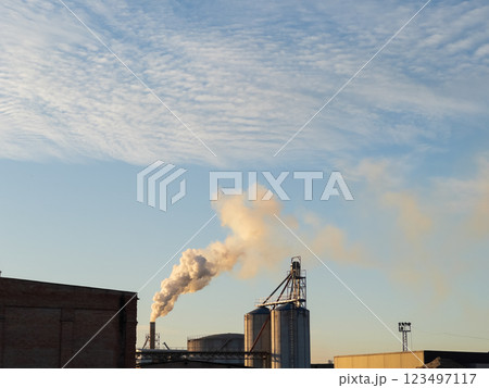 As the sun sets, smoke drifts from large industrial silos, painting a dramatic picture against the expansive blue sky. The quiet evening air hints at a bustling production nearby As the sun sets, smoke drifts from large industrial silos, painting a dramatic picture against the expansive blue sky. The quiet evening air hints at a bustling production nearby 123497117