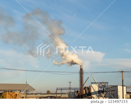 A plume of smoke rises from a tall chimney at a manufacturing plant, contrasting sharply with the clear blue sky. The sun's warm rays illuminate the scene during late afternoon A plume of smoke rises from a tall chimney at a manufacturing plant, contrasting sharply with the clear blue sky. The sun's warm rays illuminate the scene during late afternoon 123497153