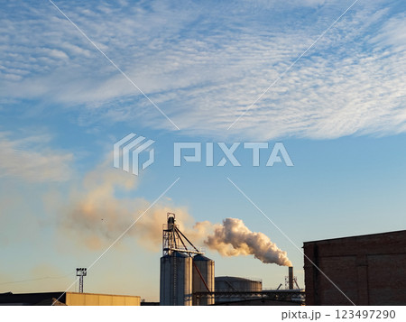 Industrial silos stand tall as smoke rises into the crisp evening air, contrasting with the bright colors of the setting sun and the textured sky above 123497290