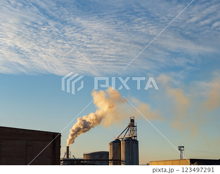 In a sprawling industrial area, smokestacks release plumes of smoke against a backdrop of fluffy clouds. The warm hues of sunset cast a golden light on the structures 123497291