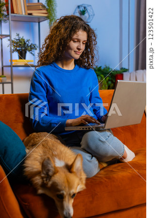Happy woman opening laptop and working remotely sitting beside corgi dog on sofa at home room 123501422