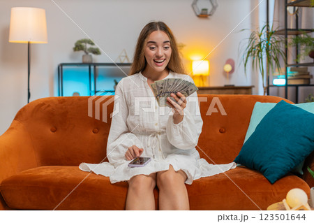Rich happy young woman counting money cash using smartphone calculator sitting on sofa at home 123501644
