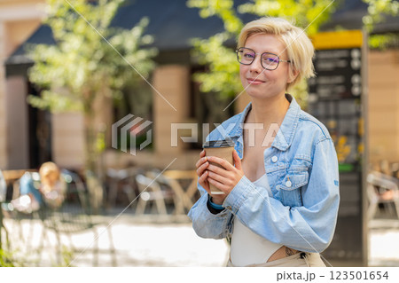 Happy Caucasian young blonde woman enjoying morning coffee hot drink and walking on city street 123501654