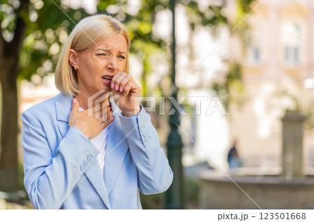 Sick mature businesswoman in formal suit suffering from sore throat while standing on city street 123501668