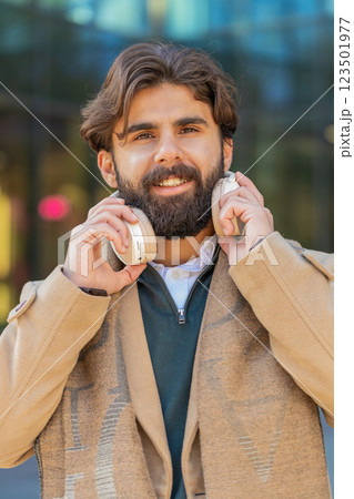 Indian young businessman taking off wireless headphones looking at camera and smiling on city street Indian young businessman taking off wireless headphones looking at camera and smiling on city street 123501977