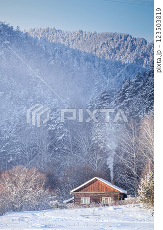 A wooden cottage for a country holiday in a winter forest against the backdrop of high mountains. Smoke from the stove is coming out of the chimney, a warm and cozy atmosphere. A Cozy Cabin Set Within A wooden cottage for a country holiday in a winter forest against the backdrop of high mountains. Smoke from the stove is coming out of the chimney, a warm and cozy atmosphere. A Cozy Cabin Set Within 123503819