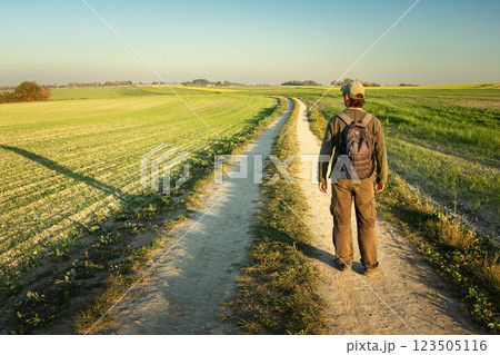 A man with a backpack stands on a dirt road and looks at rural field 123505116
