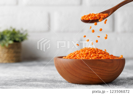 Red lentils pouring from brown wooden spoon into rustic bowl standing on grey kitchen table against white brick wall Red lentils pouring from brown wooden spoon into rustic bowl standing on grey kitchen table against white brick wall 123506872