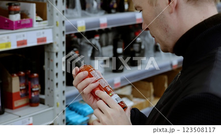 Man carefully examining hot sauce bottles in supermarket aisle, considering labels and ingredients for a well informed choice. A customer in black studies the label on a bottle of sauce Man carefully examining hot sauce bottles in supermarket aisle, considering labels and ingredients for a well informed choice. A customer in black studies the label on a bottle of sauce 123508117