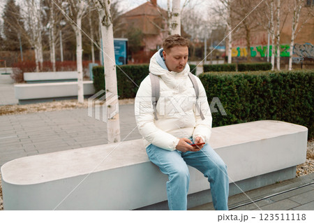 Caucasian man sitting on bench in city park using smartphone and listening music, coughing while looking at his smartphone 123511118