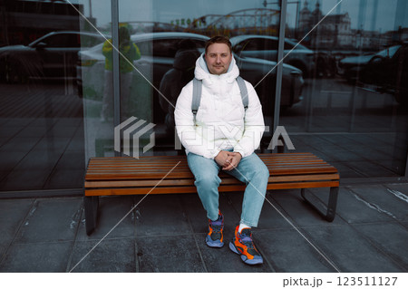 Full length of sad upset pensive thoughtful guy backpacker sit on modern bench outside in city street, enjoying break in front of modern building. Full length of sad upset pensive thoughtful guy backpacker sit on modern bench outside in city street, enjoying break in front of modern building. 123511127