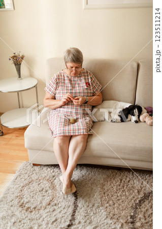 Elderly lady with glasses crochets at home while her white dog sits beside her 123511214