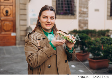 Young happy 30s woman eating sandwich on the city street. Urban food festival. Beautiful hungry girl eating tasty burger outdoor 123511380