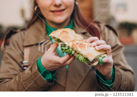 Young happy 30s woman eating sandwich on the city street. Urban food festival. Beautiful hungry girl eating tasty burger outdoor Young happy 30s woman eating sandwich on the city street. Urban food festival. Beautiful hungry girl eating tasty burger outdoor 123511381