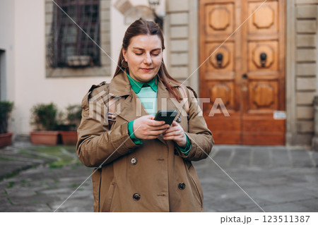 Happy cheerful young woman with backpack walking on city street checks her smartphone. Portrait of beautiful 30s girl using smartphone outdoors. Urban lifestyle concept. Happy cheerful young woman with backpack walking on city street checks her smartphone. Portrait of beautiful 30s girl using smartphone outdoors. Urban lifestyle concept. 123511387