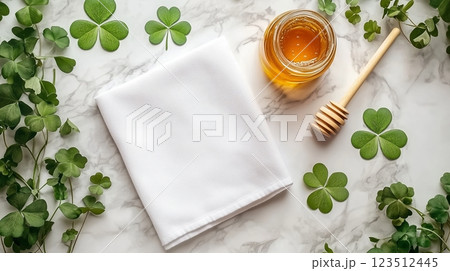 A blank white towel laying with a golden honey jar and clovers lying around it. 123512445