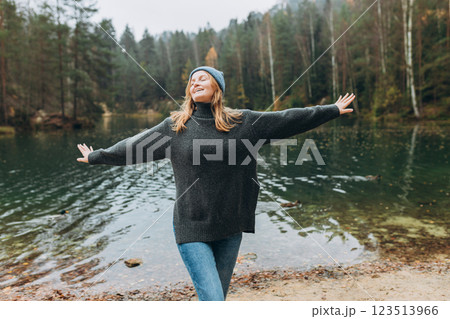Happy woman raising hands up on the banks of the lake. Young 30s woman hiker in front smiling happy, hiking in woods, warm autumn day. Happy woman raising hands up on the banks of the lake. Young 30s woman hiker in front smiling happy, hiking in woods, warm autumn day. 123513966